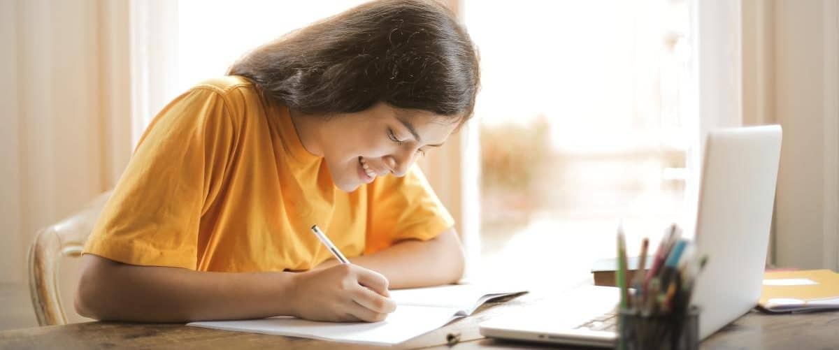 Lächelnde Studentin sitzt am Tisch und schreibt in ein Heft. Laptop und Stifte stehen vor ihr.