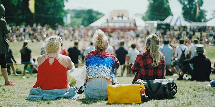 Drei junge Frauen mit dem Rücken zur Kamera sitzen auf einer Festival-Wiese - Symbolbild für Festivals in Europa.