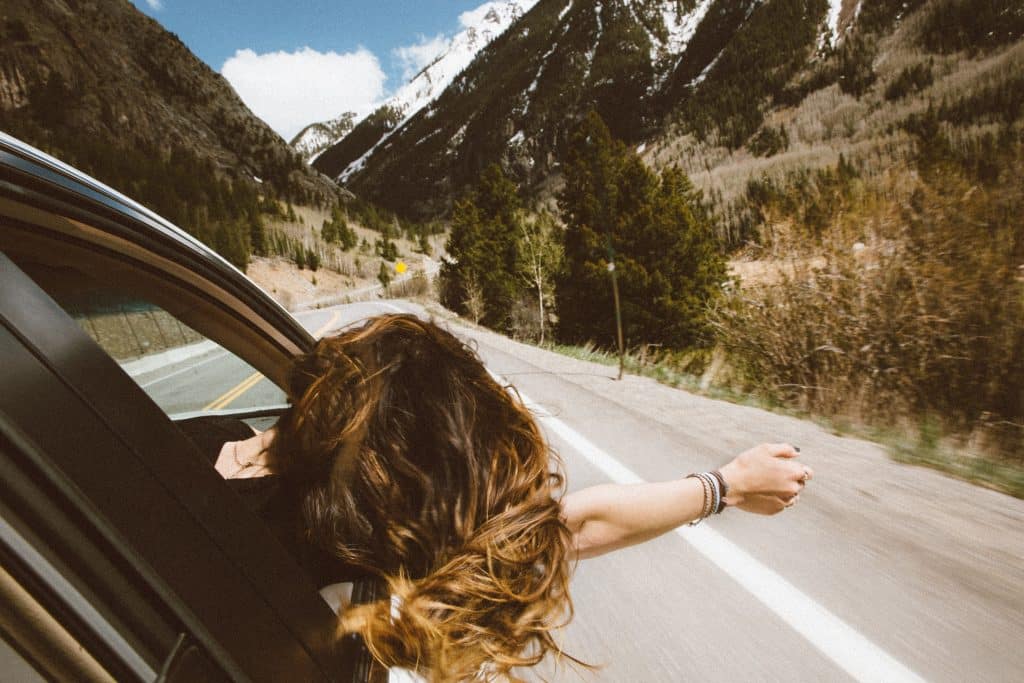 Junge Frau hält Kopf aus dem Auto während einer Fahrt auf einer einsamen Straße durch die Berge - Symbolbild für Mietwagen und Carsharing