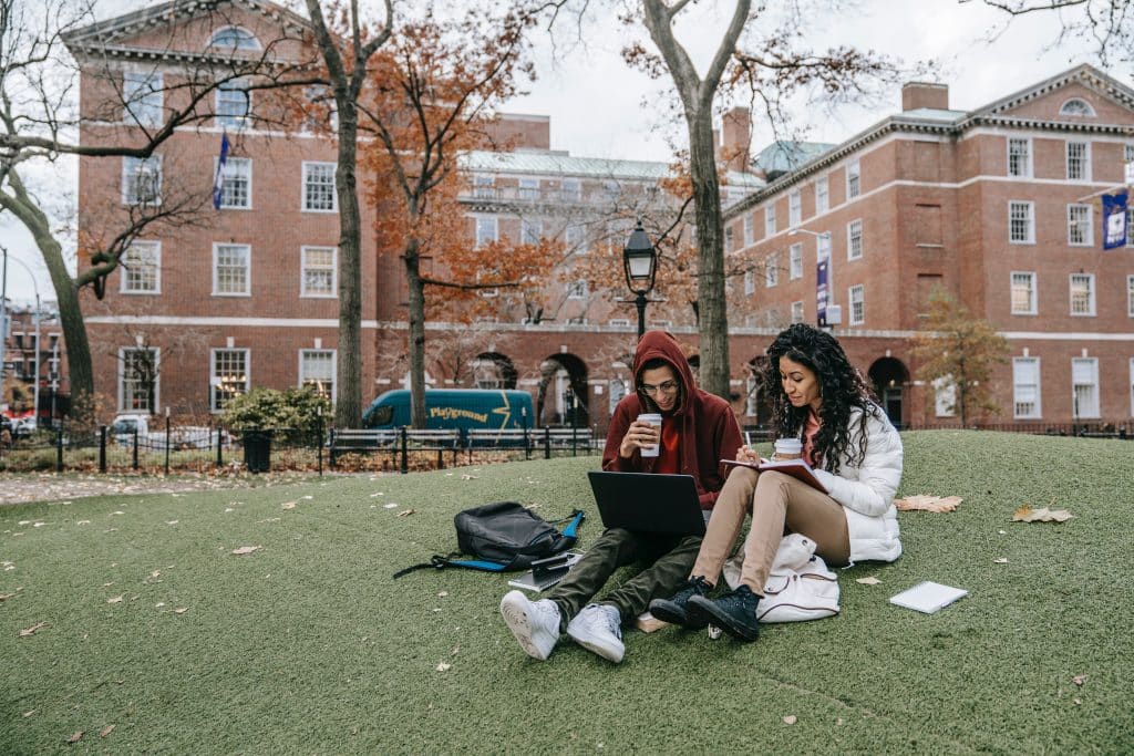 Junger Mann und junge Frau sitzen gemeinsam auf dem Rasen vor Universität, schauen auf einen Laptop und trinken Kaffee - Symbolbild für Studieren im Ausland
