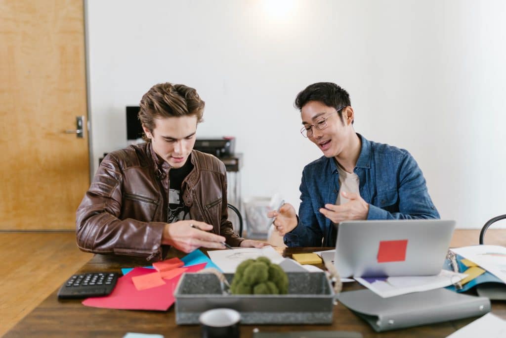 Zwei junge Männer sitzen an einem Schreibtisch mit Laptop vor sich und diskutieren - Symbolbild für Existenzgründung