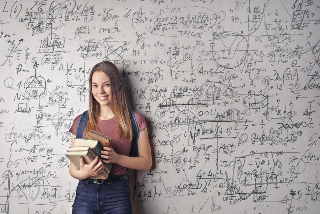 Junge Frau mit Büchern in der Hand steht vor einer Wand voller Formeln - Symbolbild zum Studienbeginn