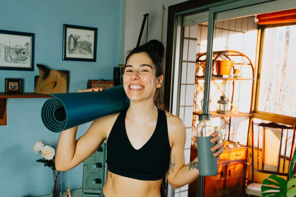 Eine glückliche junge Frau in Sportklamotten mit Yoga-Matte und Trinkflasche - Symbolbild für Sport im Studium.
