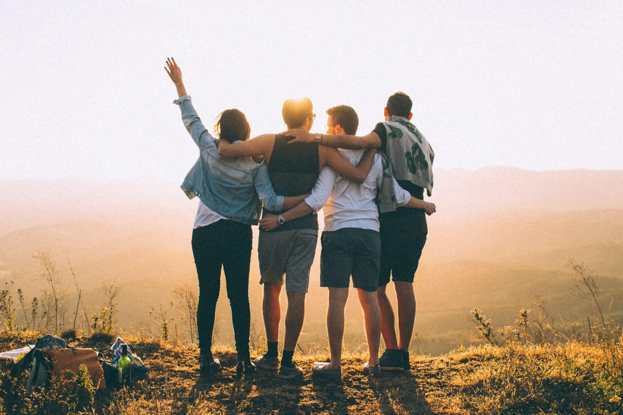 Vier Freunde mit dem Rücken zur Kamera stehen Arm in Arm vor einer hügeligen Landschaft - Symbolbild für einen Kurzurlaub im Semester.