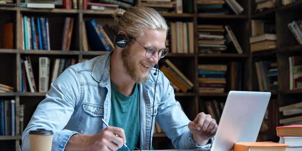 Blonder Mann mit Kopfhörern sitzt lächelnd vor seinem Laptop.