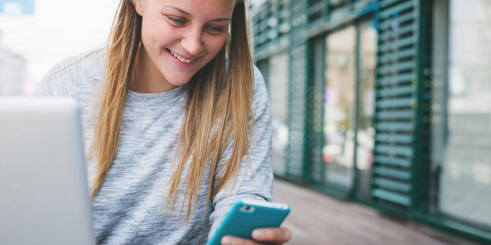 Blonde junge Frau sitzt vor einem Laptop und schaut lächelnd auf ihr Smartphone.