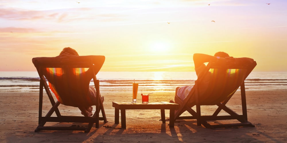 Zwei Menschen entspannen auf Liegestühlen amf Strand mit dem Blick zum Sonnenuntergang.