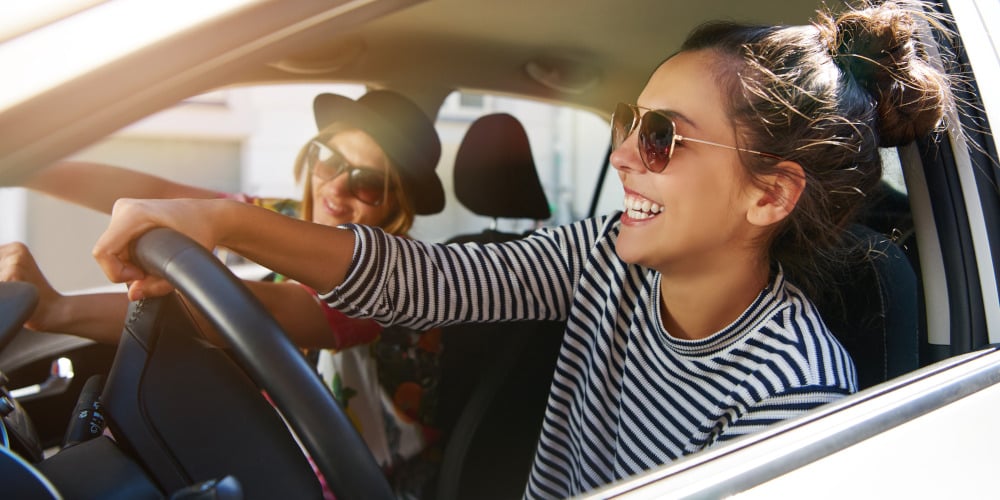 Zwei junge Frauen mit Sonnenbrillen fahren lachend und lächelnd in einem Auto in der Stadt, Blick durch das offene Seitenfenster.